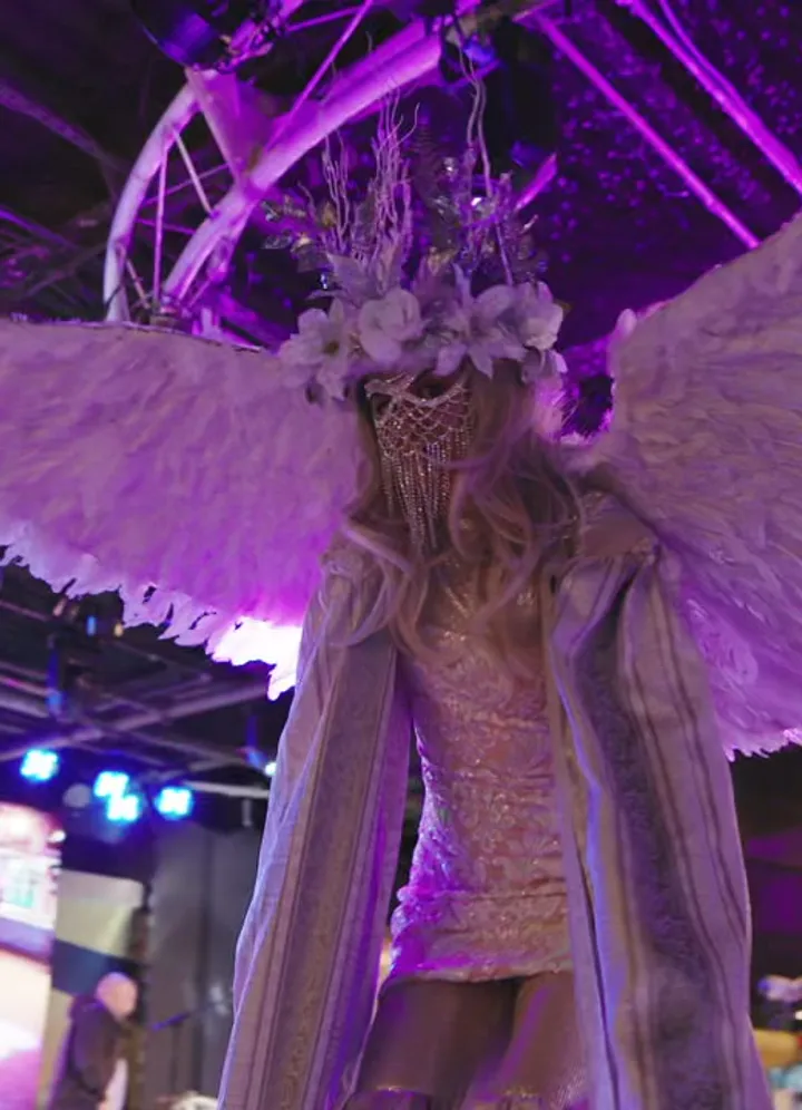 A performer in a white feathered angel-wing costume on a lit stage